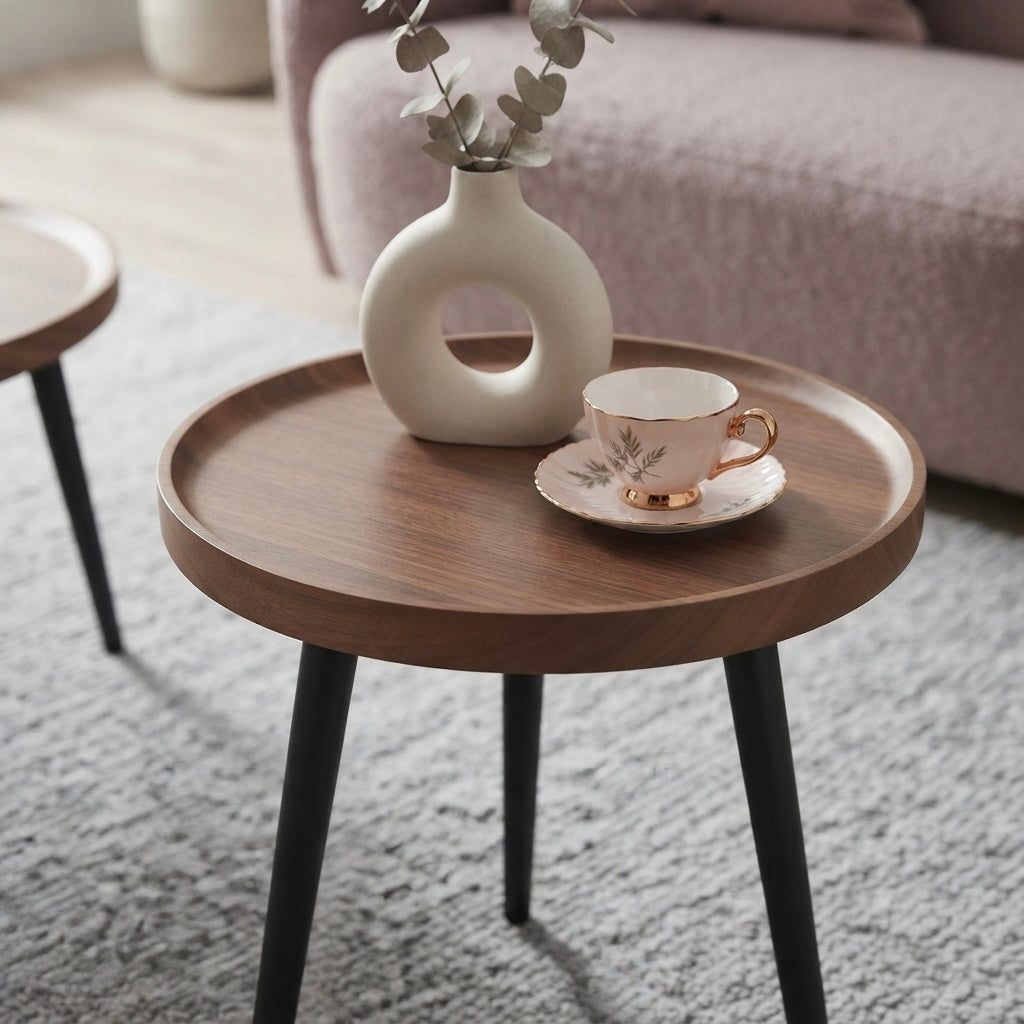 A round wooden side table with black metal legs in a living room setting, styled with a white ceramic vase and a white teacup on a saucer.
